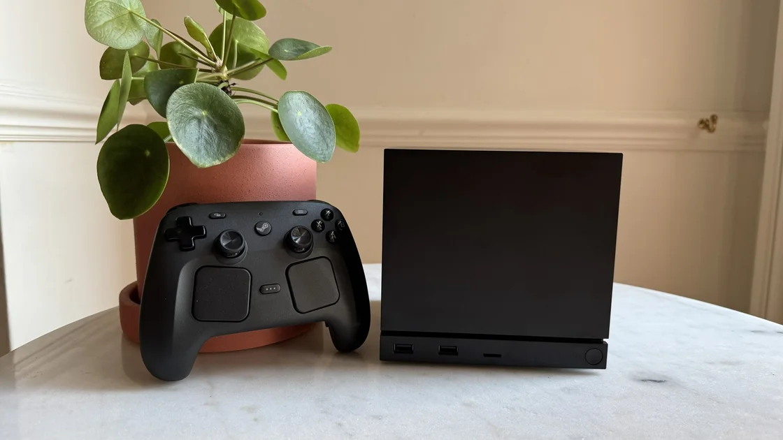 Steam Machine photograph showing the steam machine with black faceplate on a marble circular table in front of a grey/beige wall, with a plant pot and the Steam Controller next to it