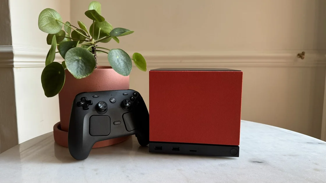 Steam Machine photograph showing the steam machine with red faceplate on a marble circular table in front of a grey/beige wall, with a plant pot and the Steam Controller next to it